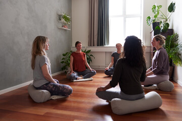A group of women sit on a cushion on a floor. They sit still with their eyes closed and meditate in a circle during yoga class.
