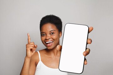 Pointing up healthy woman holding smartphone with white empty blank screen display on gray studio...