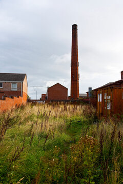 A single tall industrial brick chimney stands out against a cloudy sky in a grassy urban field