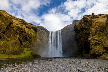 A majestic Skogafoss waterfall cascades down through the rocky mountains on a bright sunny day,...