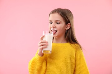 Funny little girl with milk mustache holding glass of tasty dairy drink on pink background