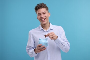 Happy man putting coins into piggy bank on light blue background