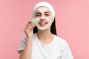 Young woman with headband washing her face using sponge on pink background