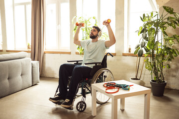 Young man sitting in wheelchair in headphones, training, doing exercises with dumbbells at home....