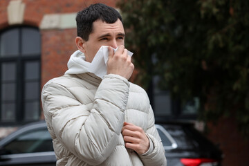 Sick young man with tissue blowing runny nose outdoors. Cold symptom