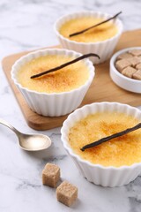 Delicious creme brulee in bowls, vanilla pods, sugar cubes and spoon on white marble table