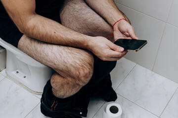 A man is sitting on the toilet with a phone in his hand. Guy's morning routine