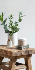 A wooden stool table against a white wall and there are some cosmetic products and bottles on it an a white vase with plant inside. web banner style.