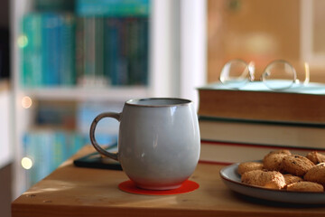 Cup of tea or coffee, pile of books, plate of cookies, reading glasses, e-reader and pen on the table. Colorful rainbow bookshelf in the background. Selective focus.