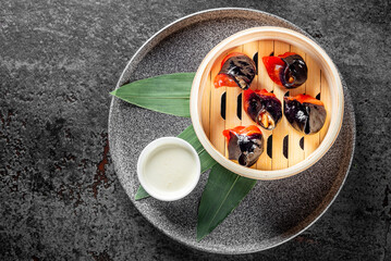 Top view of a bamboo steamer with dim sum, accompanied by green leaves and a cup of sauce on a textured surface