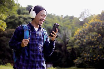 Asian man in a casual outfit, sporting a beanie, plaid shirt, and headphones, checks his phone with...