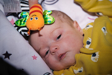Close-up of a newborn baby lying down with a colorful toy, wearing a yellow onesie with animal...
