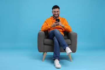Happy young man using smartphone on armchair against light blue background
