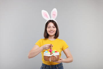 Easter celebration. Happy woman with bunny ears and wicker basket full of painted eggs on grey background