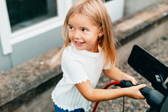 Smiling blond girl plugging charger in electric car at front yard