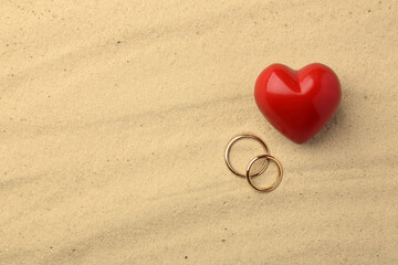 Fototapeta na wymiar Honeymoon concept. Two golden rings and red heart on sand, top view. Space for text