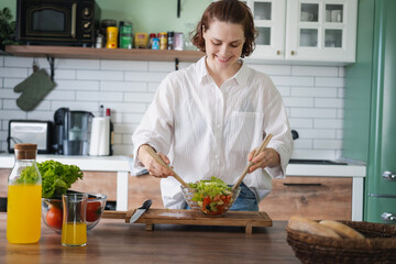 Young Caucasian woman cooking salad from green fresh vegetables while standing in the kitchen at home - 780430595
