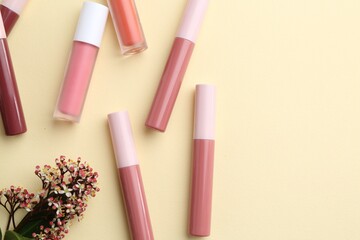 Different lip glosses and flowers on pale yellow background, flat lay. Space for text