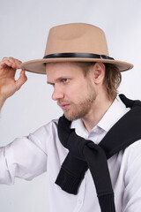 Close-up shot of a young blond-haired man in a white shirt and a beige felt fedora hat with a black strap. A black sweater is draped over a man's shoulders. The man in the hat is posing on a light bac