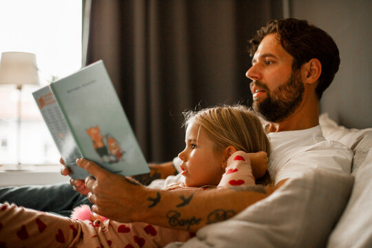 Father and daughter reading picture book while reclining on bed at home