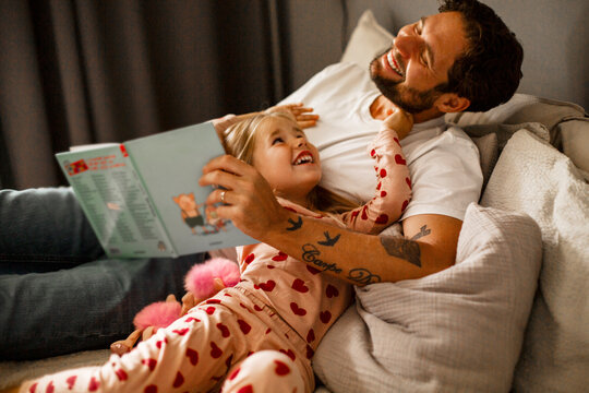 Cheerful father and daughter reclining with picture book on bed at home