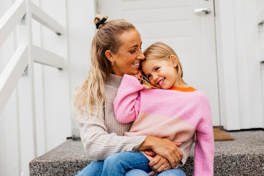 Happy blond woman sitting on front stoop with daughter in lap