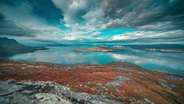 Stormy clouds whirl above the calm mirrorlike fjord and autumn tundra in the timelapse video.