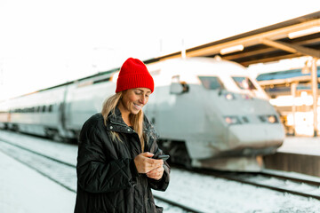 Smiling woman text messaging on mobile phone while standing at railroad station