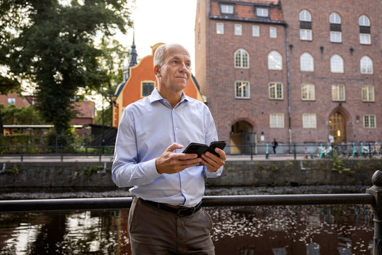Senior man holding smart phone and standing near river in front of buildings