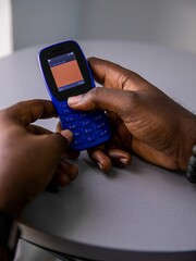 Vertical shot of a dark-skinned person holding a feature phone with the background of a white table