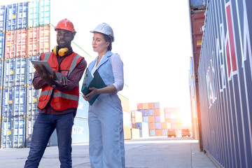 Two workers with safety helmet at logistic shipping cargo containers yard. African engineer man using digital tablet to report results to beautiful woman boss who holds document folder at workplace.