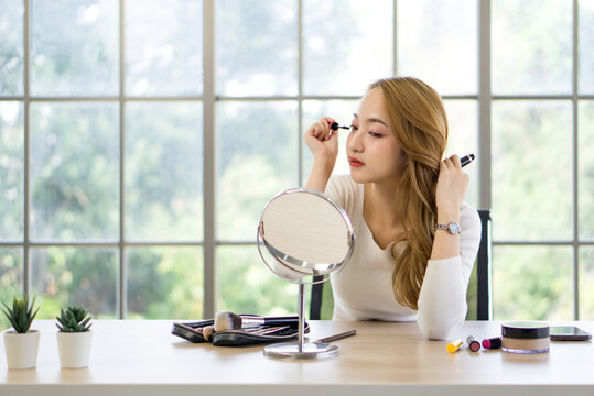 A woman is applying makeup while looking into a round mirror on a table. She is focused on applying mascara and has a collection of makeup items and brushes in front of her as well as a makeup palette