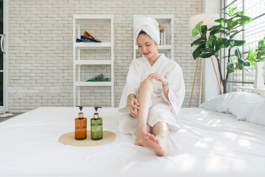 Young asian woman in a white bathrobe and towel on head, sit on a bed with lotion bottle nearby. Spreading body lotion cream gel on leg and knee after showering and taking bath, healthcare