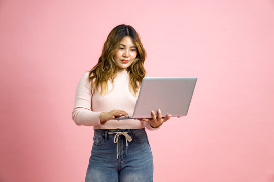 Young asian woman in casual clothing, holding and looking at a laptop computer. Portrait on a simple pink background with studio light.