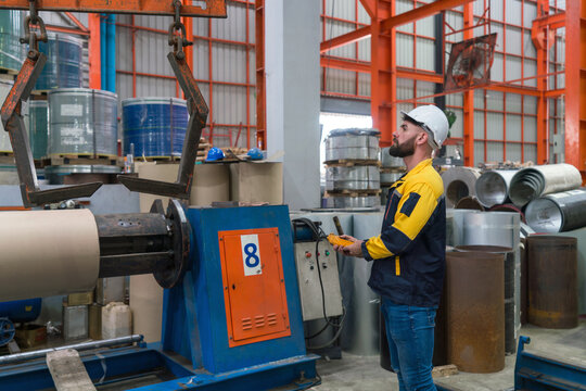 Young technician wearing a hardhat and a high-visibility jacket with safety color, inspecting machinery in industrial plant. A busy industrial environment with machinery in production line.