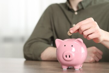 Financial savings. Man putting coin into piggy bank at wooden table, closeup