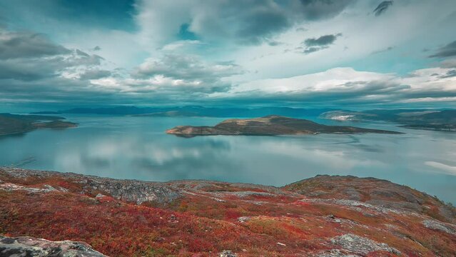 Stormy clouds backlit by the autumn sun whirl above the calm fjord and mountains in the timelapse video.