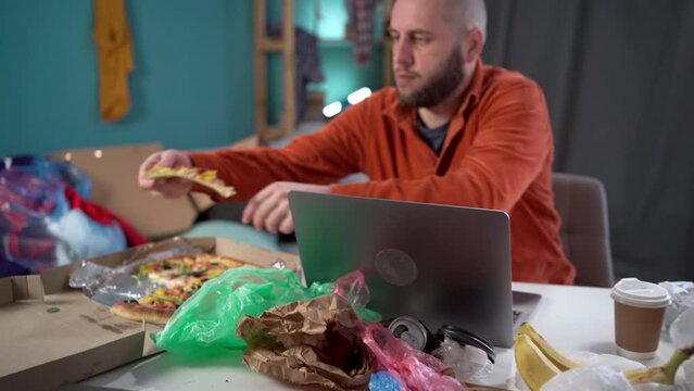 mess, disorder and chaos concept. Male freelancer eating pizza working in messy home living room with scattered stuff