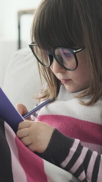 Portrait of a preschool Caucasian focused girl drawing or writing in notebook on sofa at home. Learning or imagination concept. Vertical video.