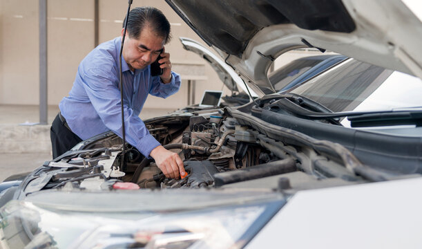 Senior man work on a vehicle, engaged in a car repair or inspection while using a mobile phone, possibly seeking advice or discussing the issue with someone, focus on the engine compartment of the car