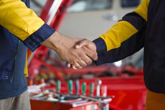 Two mechanic in work uniform engaged in a handshake. Atmosphere in automotive workshop or garage with hand tools laid on the tool rack.