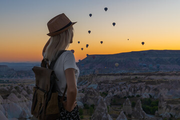 Young woman with a craft backpack in a hat admires the sunrise and flying balloons in Cappadocia,...