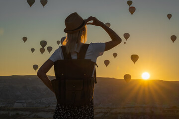 Young female tourist in a hat with a backpack admires the amazing scenery of hot air balloon...