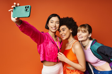 Three women, representing different cultures, enjoying a playful moment as they take a selfie with a cell phone against an orange background.