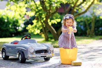 Cute gorgeous toddler girl washing big old toy car in summer garden, outdoors. Happy healthy little...