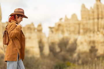 A woman wearing a brown jacket and a brown hat is walking in a desert. She is carrying a backpack...