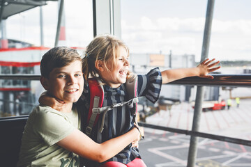 Little girl and school boy at the airport waiting for boarding at big window. Two kids stands at window against the backdrop of airplanes. Happy children, siblings leaving for family summer vacation