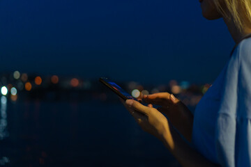 Close-up of a woman's hands with a mobile phone on the background of a blurred city with lights, water, copy space.