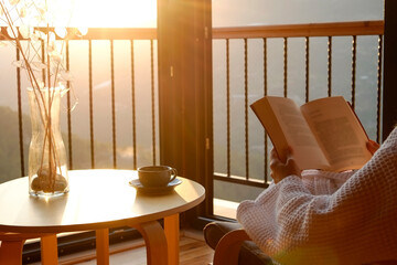 Cropped shot of unrecognizable woman in a robe enjoying her evening read by the window at sunset. Cup of tea on the table. Close up, copy space, background.