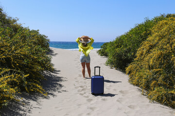 Back view of a young woman entering the beach with a suitcase. The beginning of vacation at tropical destination. Copy space, background.